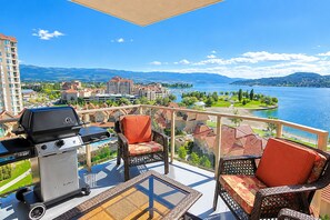A balcony with outdoor furniture, a grill, and a view of a lake and mountains.