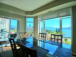 A dining area with a glass table and chairs, large windows with a view of a body of water and buildings. - Sunset Waterfront Resort 9. Luxury Lakeside, Stunning 3 bed/2bath Kelowna Condo. (Kelowna)