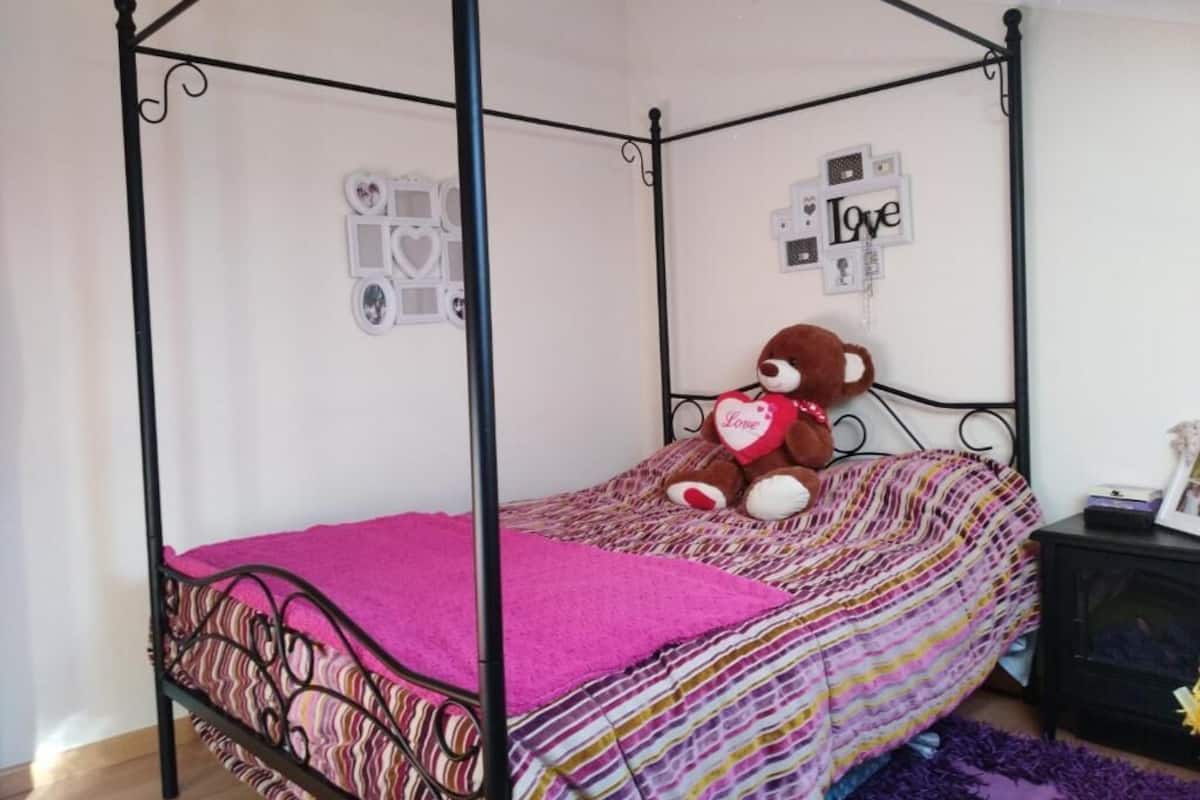 A four-poster bed with a pink blanket, a teddy bear with a heart, and wall decorations.