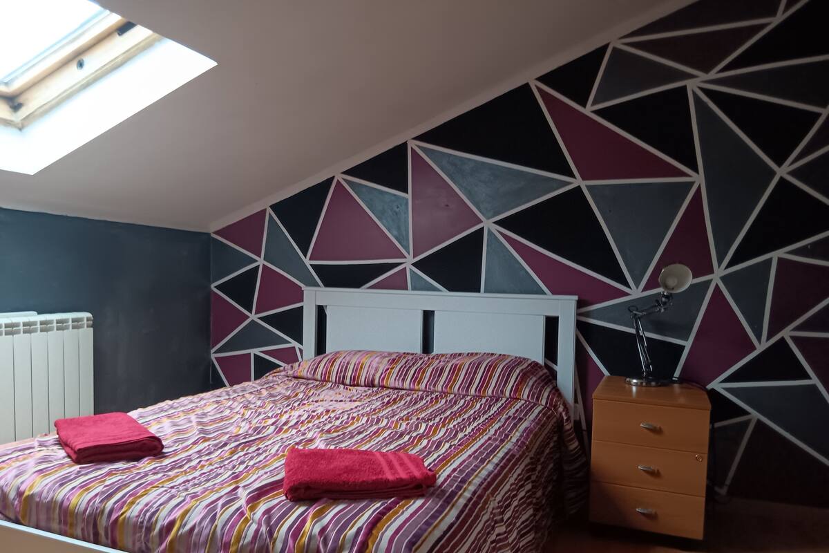 A bedroom with a geometric-patterned wall, a bed with striped bedding, a red towel, and a nightstand with a lamp.