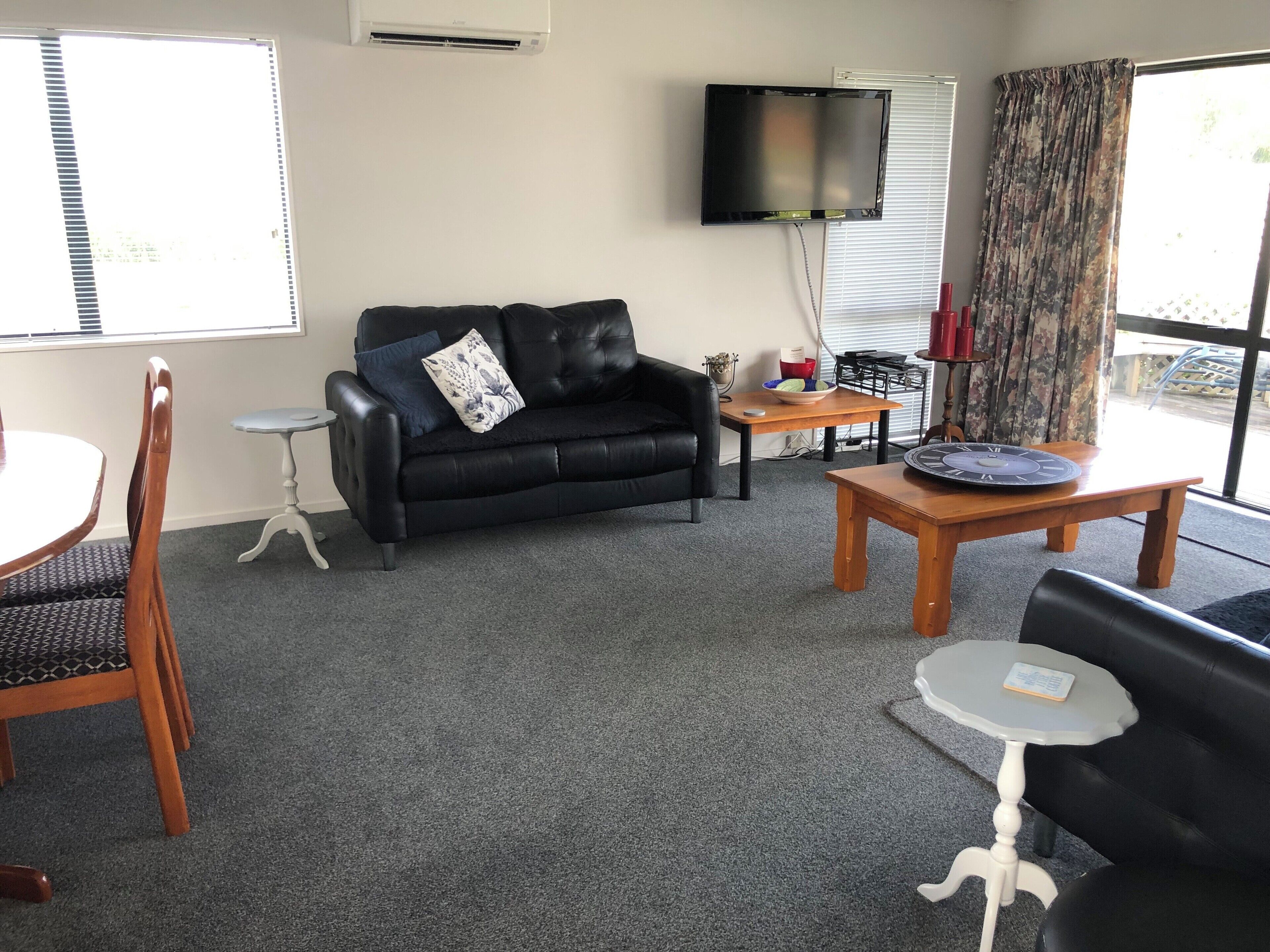 Uma sala de estar com um sofá de couro preto, uma mesa de centro de madeira, uma pequena mesa lateral branca, uma mesa de jantar com cadeiras e uma TV de tela plana fixada na parede.