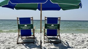 To strandstole under en blå og grøn parasol med skilt fra 'Sandpiper Cove Chair Rentals', placeret på en sandstrand med havet i baggrunden.