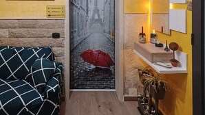 Habitació d'hotel amb una porta que mostra un carrer plujós amb la Torre Eiffel, un sofà quadriculat, una petita estant amb productes de tocador i una planta en test.