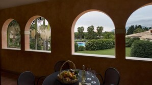 A dining area with a table set for two, a fruit basket, and a view of a landscaped garden with a pool. - Tranquil Lantana Resort 2 Bedroom Apartments Sleeps 7 Persons (Pula)