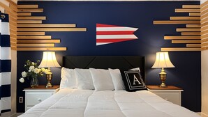 A neatly made bed with white bedding, flanked by bedside tables with lamps and a vase of flowers. A decorative wall piece with a flag design is centered above the bed. - Acadia Ocean View Hotel (Bar Harbor)