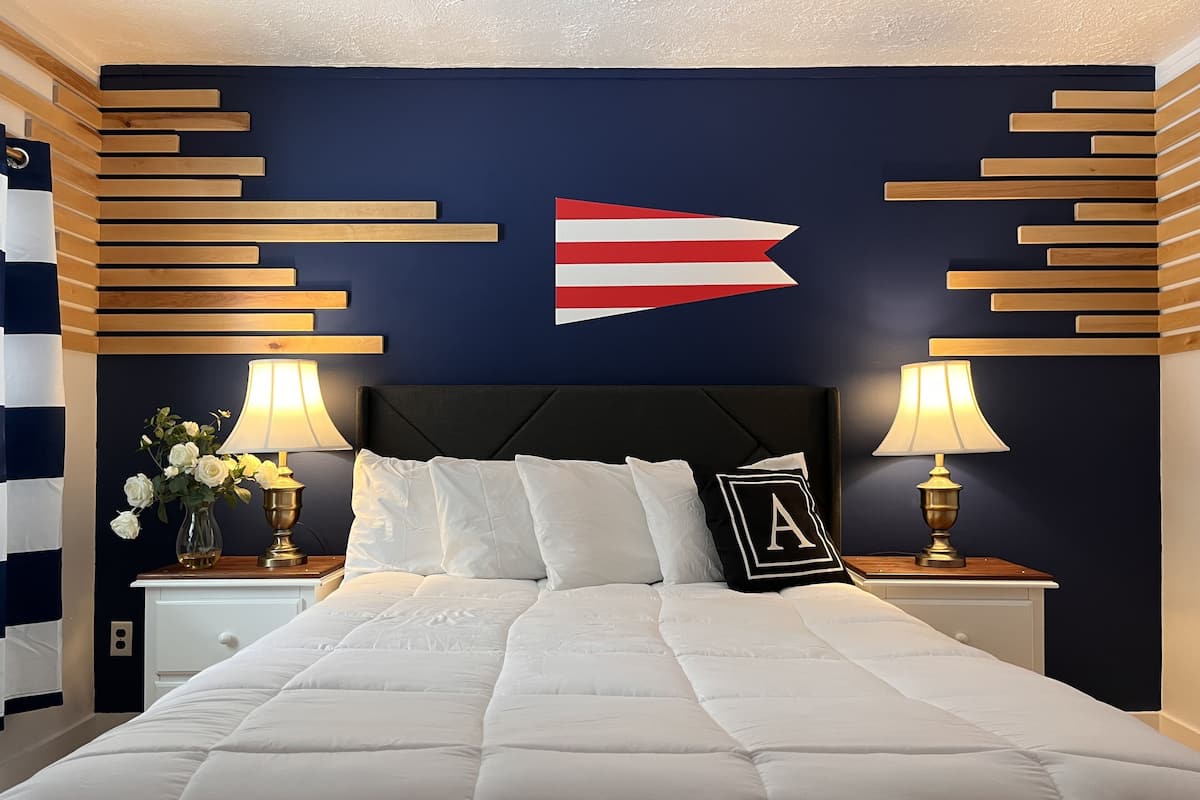 A neatly made bed with white bedding, flanked by bedside tables with lamps and a vase of flowers. A decorative wall piece with a flag design is centered above the bed.