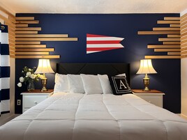 Una cama bien tendida con ropa blanca, flanqueada por mesitas de noche con lámparas y un jarrón de flores. Una pieza decorativa con diseño de bandera está centrada sobre la cama.
