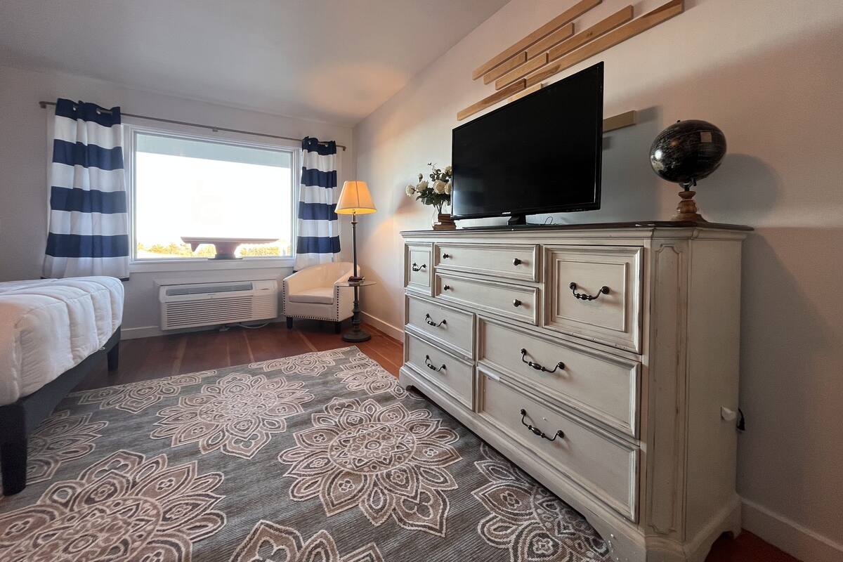 A bedroom with a bed, a dresser, a television, and a window with curtains.