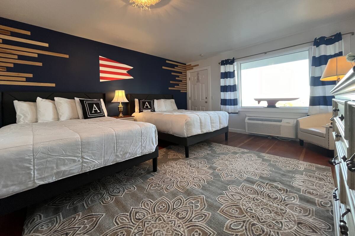 A hotel room with two beds, a patterned rug, a ceiling light, a window with curtains, and a stove.