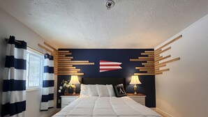 A bedroom with a large bed, a nightstand with a lamp, a wall-mounted flag, and a window with curtains. - Acadia Ocean View Hotel (Bar Harbor)