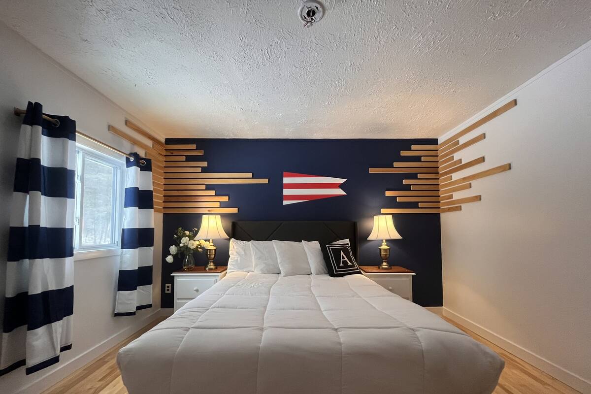 A bedroom with a large bed, a nightstand with a lamp, a wall-mounted flag, and a window with curtains.