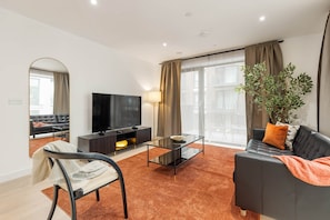 A modern living room with a sofa, a TV, a glass coffee table, and a large window with curtains.