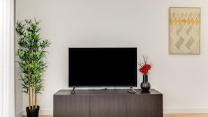 A modern living room with a flat-screen TV on a dark wood cabinet, a tall bamboo plant, and a vase with red flowers on a small table. - Immerse yourself in our contemporary East London 2-bedroom, 2-bathroom apartment (London)