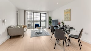 A modern living room with a sofa, a dining table with chairs, a coffee table, and a large window with blinds. - Immerse yourself in our contemporary East London 2-bedroom, 2-bathroom apartment (London)