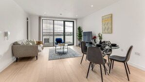 A modern living room with a sofa, a dining table with chairs, a coffee table, and a large window with blinds. - The Water - Aqua Apt (London)