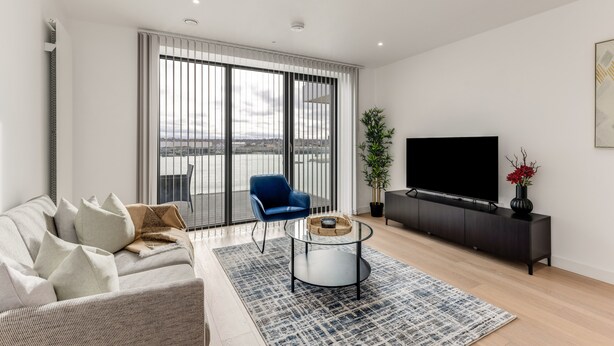 A modern living room with a sofa, a blue chair, a coffee table, a TV on a black stand, and a large window with a view. - The Water - Aqua Apt (London)