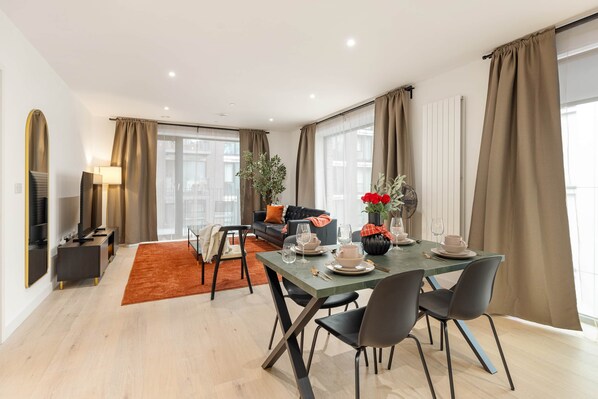 A modern living room with a dining area, large windows, and a view of the city. - The Crow - 1 Bedroom Apt (London)