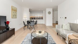A modern living room with a flat-screen TV, a dining area with a table and chairs, and a kitchen in the background. - The Water - Aqua Apt (London)