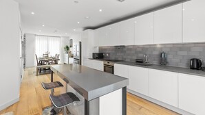 Een moderne keuken met witte kastjes, een grijze aanrechtblad en roestvrijstalen apparatuur. Er is een eethoek met een houten tafel en stoelen.