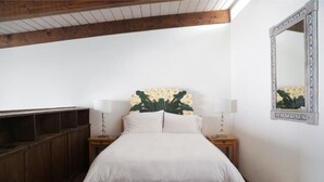 Un dormitorio con cama de madera, ropa de cama blanca, dos mesitas de noche con lámparas, un espejo y un banco de madera a lo largo de la pared.