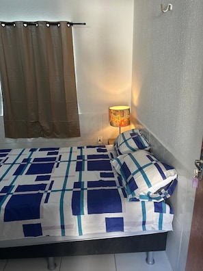 A small, neatly made bed with a blue and white checkered bedspread and matching pillows, a bedside lamp with a floral lampshade, and a brown curtain on a window. - Grand Maracanaú (Maracanau)