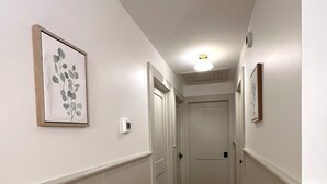 A narrow hallway with wooden flooring, white walls, and a ceiling light. There are framed pictures on the walls and a door at the end of the hallway. - Quaint Village Cottage (Hartwick)