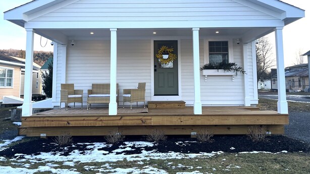 A white single-story house with a front porch, a green door, and a wreath. - Quaint Village Cottage (Hartwick)