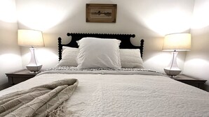 A neatly made bed with white linens and a beige throw blanket, flanked by two bedside tables with lamps, against a plain wall with a framed picture. - Quaint Village Cottage (Hartwick)