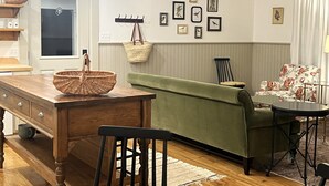 A dining area with a wooden table, a green sofa, and a chair. There are wall-mounted picture frames and a ceiling fan. - Quaint Village Cottage (Hartwick)