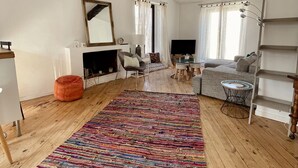 Een ruime woonkamer met een groot, kleurrijk vloerkleed, een open haard, een televisie en een gezellige zithoek.