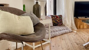 Een knusse woonkamer met een bruine leren fauteuil, een witte tafel met een lamp, een televisie en een raam met transparante gordijnen.