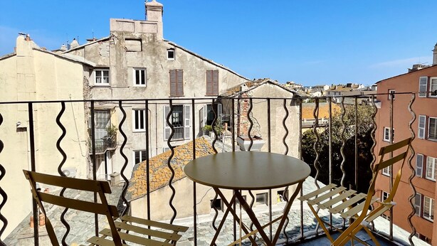 Een terras met een ronde tafel en twee stoelen, met uitzicht op een stadsgezicht met gebouwen en een heldere blauwe lucht.