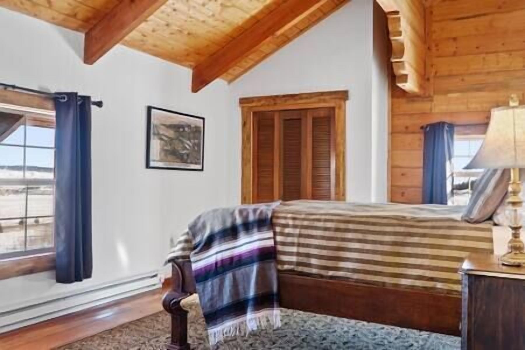 Ein Schlafzimmer mit einem Bett, einem Fenster mit Vorhängen, einer Holzbalkendecke und einem gerahmten Bild an der Wand.