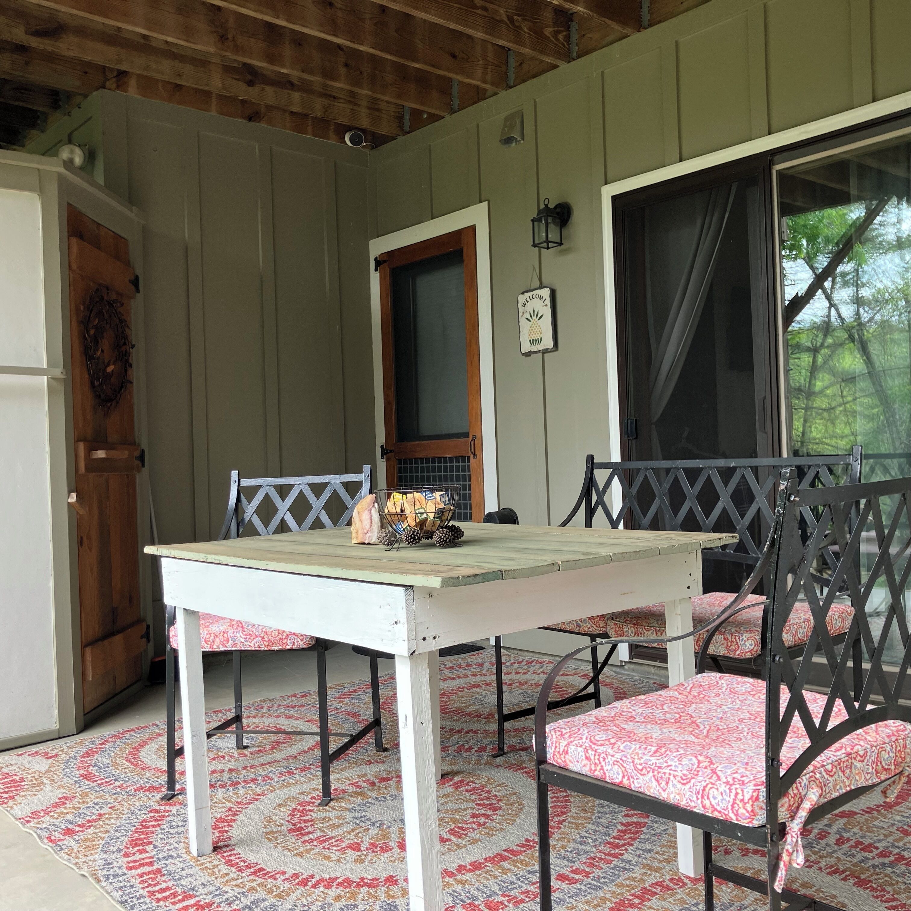 A covered porch with a table and chairs, a wooden door, a welcome sign, and a window with curtains.