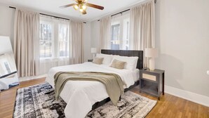 Un dormitorio con cama, mesita de noche, lámpara, ventilador de techo y ventanas amplias con cortinas.