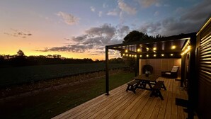A wooden deck with a pergola and string lights, overlooking a field and a body of water. - Modern 2 Bedroom, 2 Bathroom Cottage with Farm Views (Bombay)