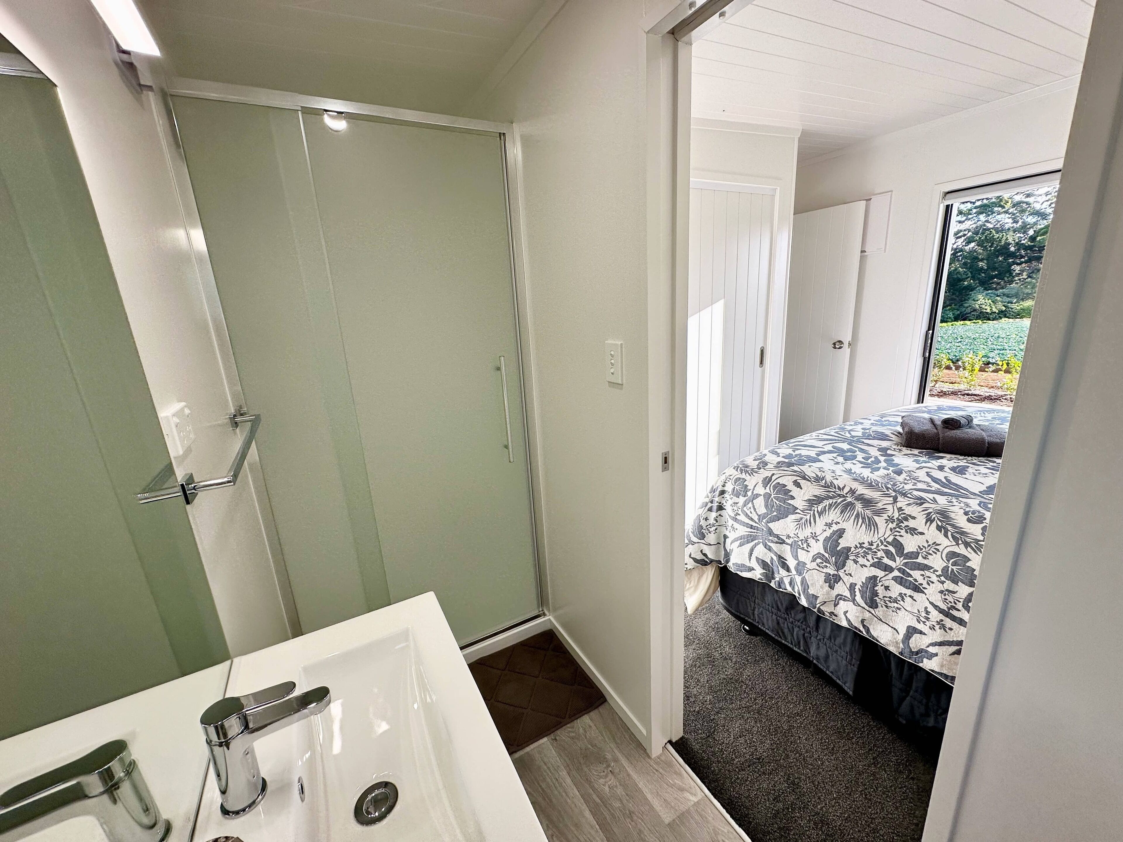 Bagno con lavandino bianco, doccia e porta che dà su una camera da letto con letto e vista sul verde all'esterno.