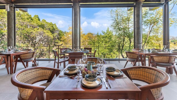 Un área de comedor al aire libre con mesas preparadas para dos, rodeada de sillas de mimbre y vistas a un paisaje boscoso.