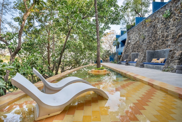 Un área de piscina con una estructura blanca curva, un muro de piedra y una zona de estar con cojines.