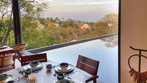 Mesa de comedor dispuesta con platos, tazones y cubiertos, con vistas a un paisaje que incluye un puente y edificios.