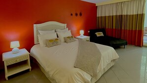A hotel room with a large bed, a nightstand with a lamp, a chair, and a window with curtains. - Résidence de la vieille tour  (Le Gosier)