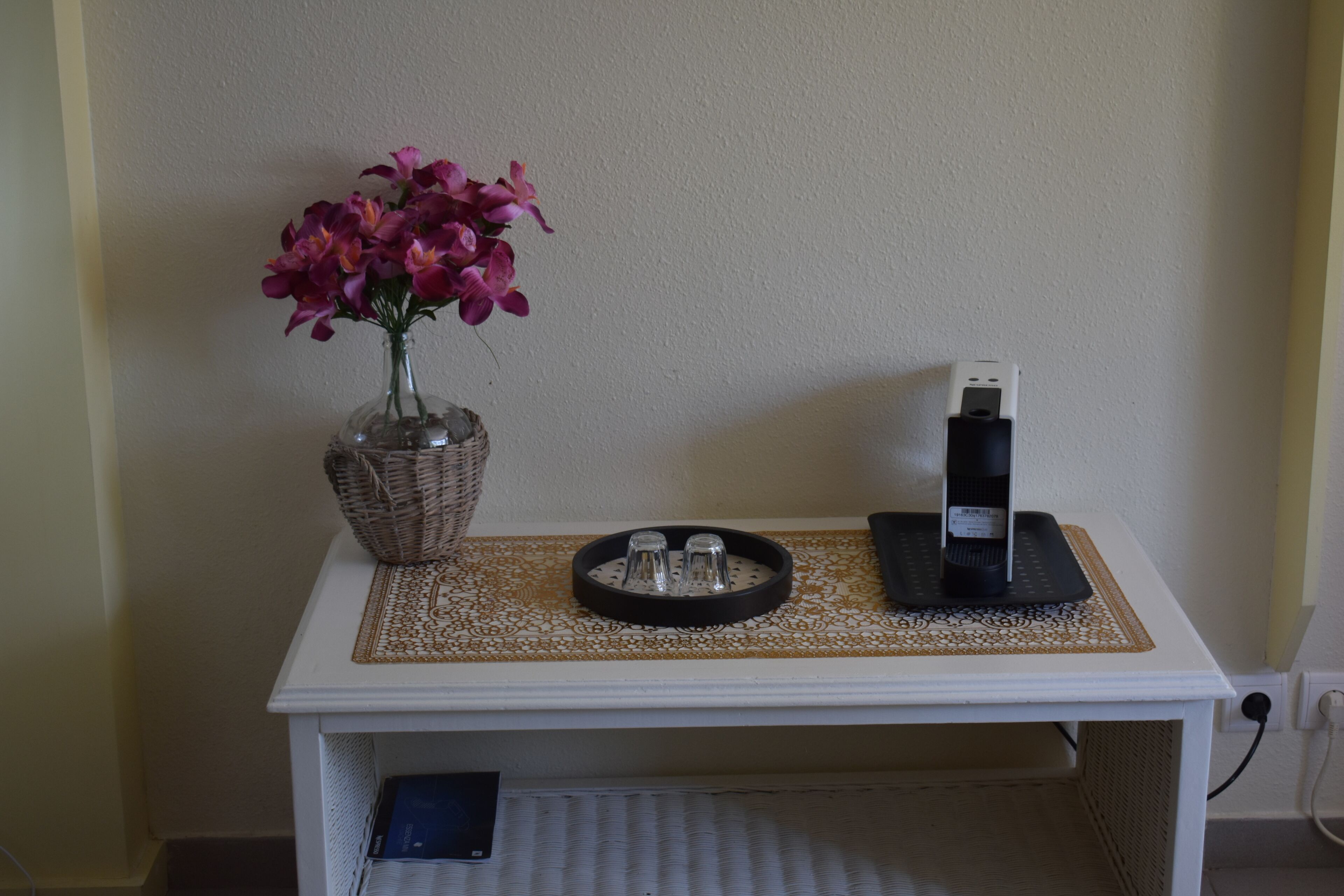 A small white table with a vase of purple flowers, a black phone charger, and a woven basket.