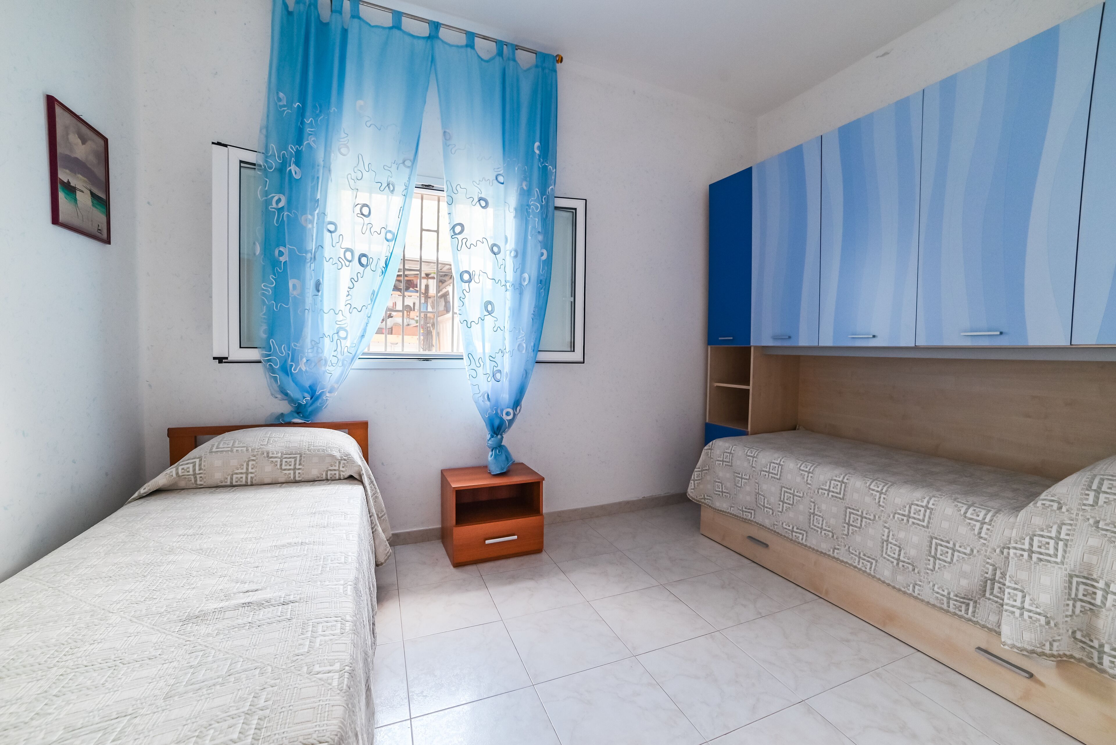 A bedroom with two beds, a window with blue curtains, a small wooden nightstand, and a wardrobe with blue panels.