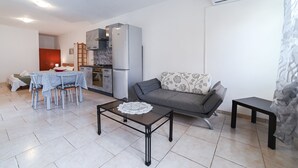 A modern living room with a glass coffee table, a grey sofa, a dining table with chairs, a kitchen area with a refrigerator, and a small black table.