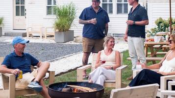 A group of people seated around a fire pit in an outdoor setting with wooden chairs and a white building in the background.