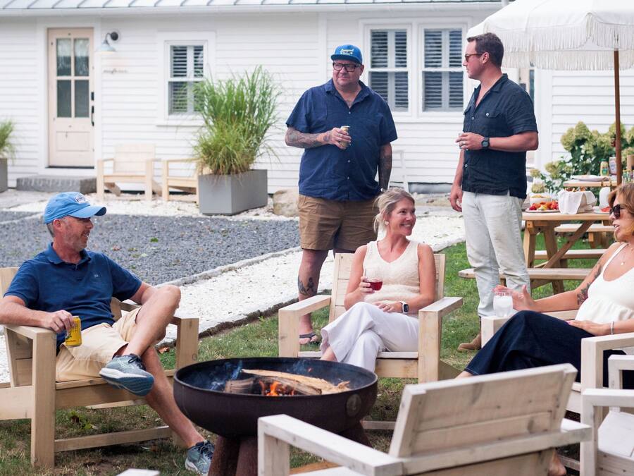 A group of people seated around a fire pit in an outdoor setting with wooden chairs and a white building in the background.