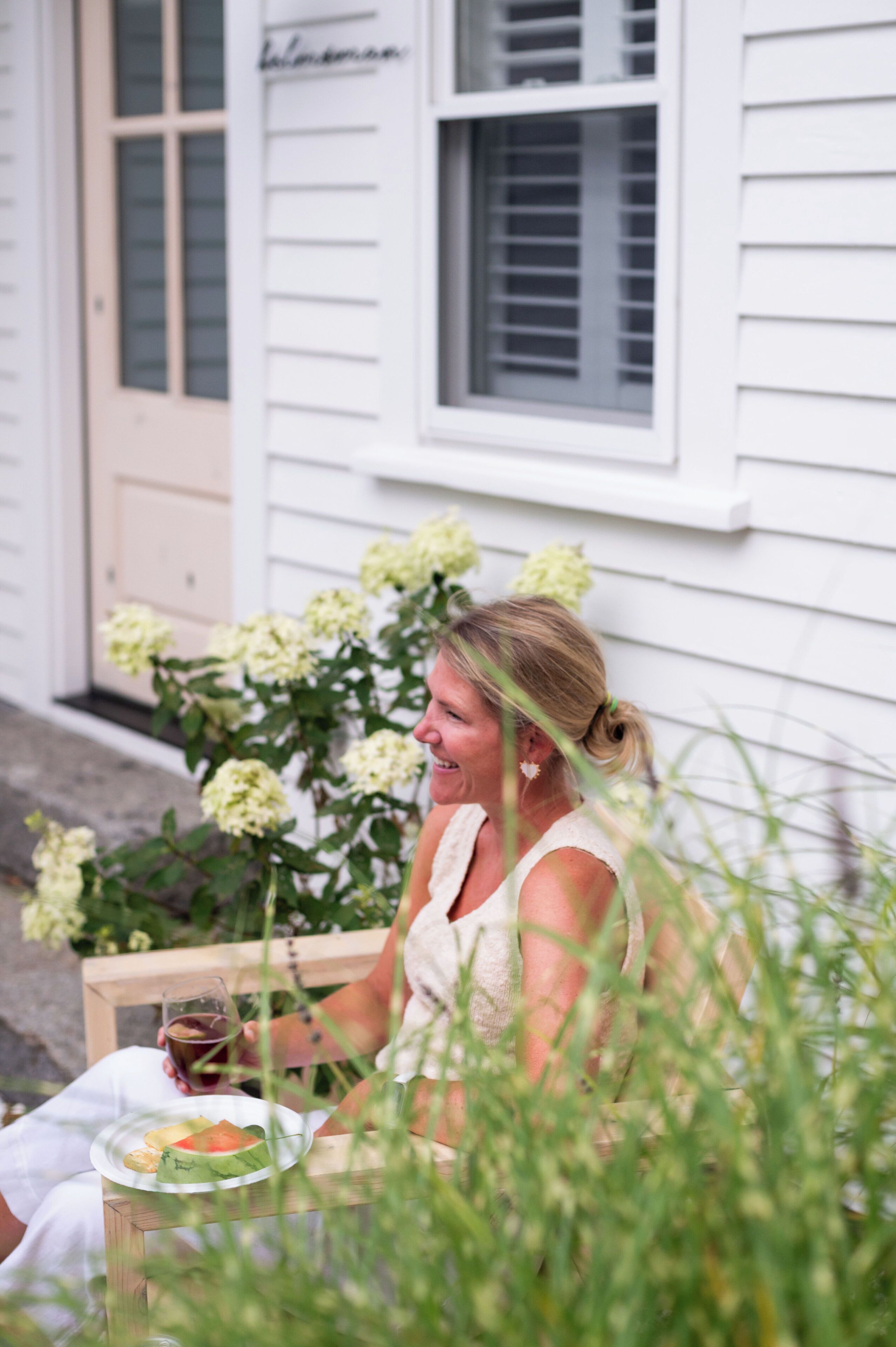 A person sitting on a bench outside a house with a plate of food and a drink.