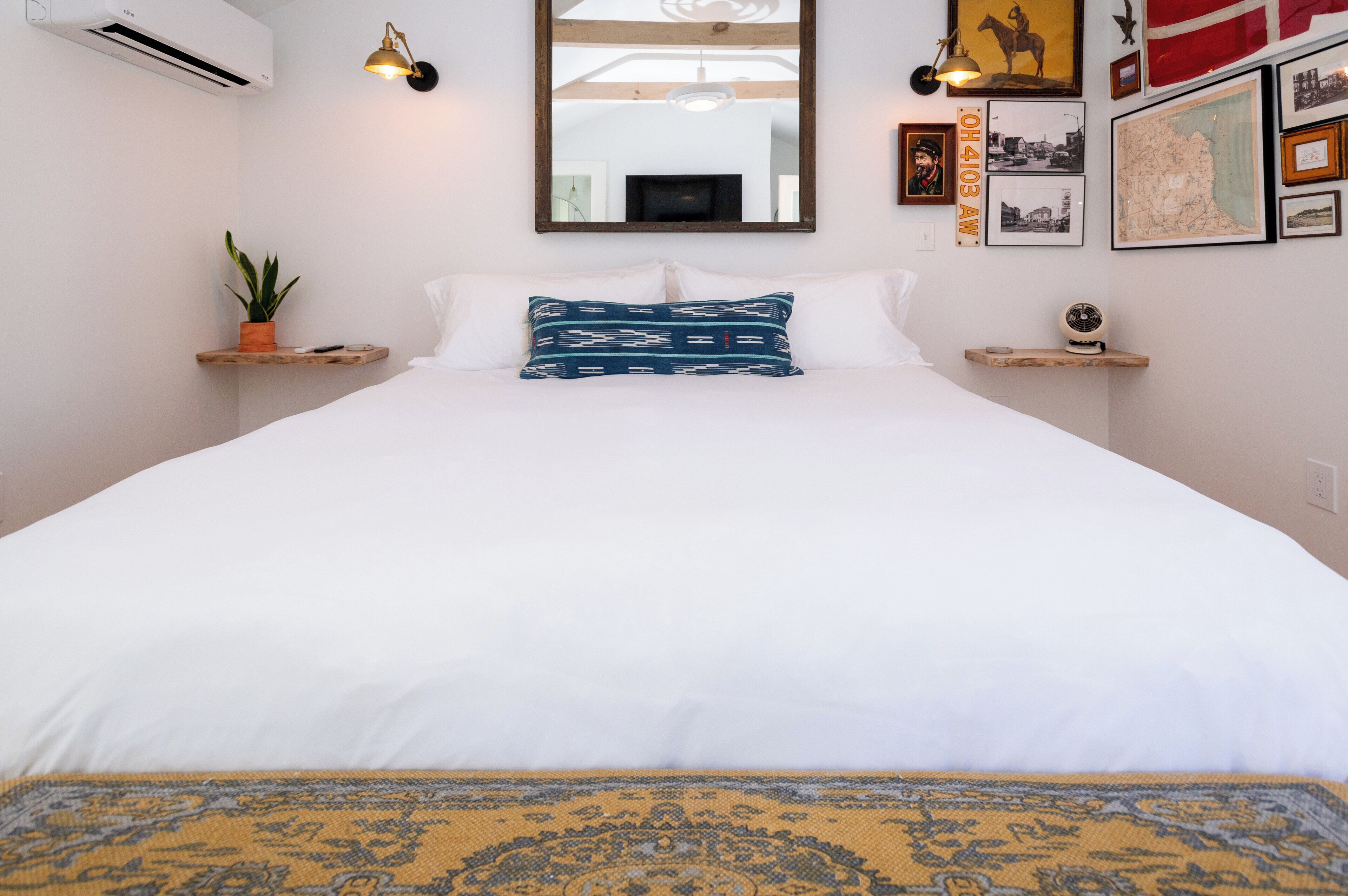 A neatly made bed with white linens, a blue patterned pillow, and a yellow patterned rug. There are two bedside tables with small lamps, a potted plant, and a wall-mounted TV.