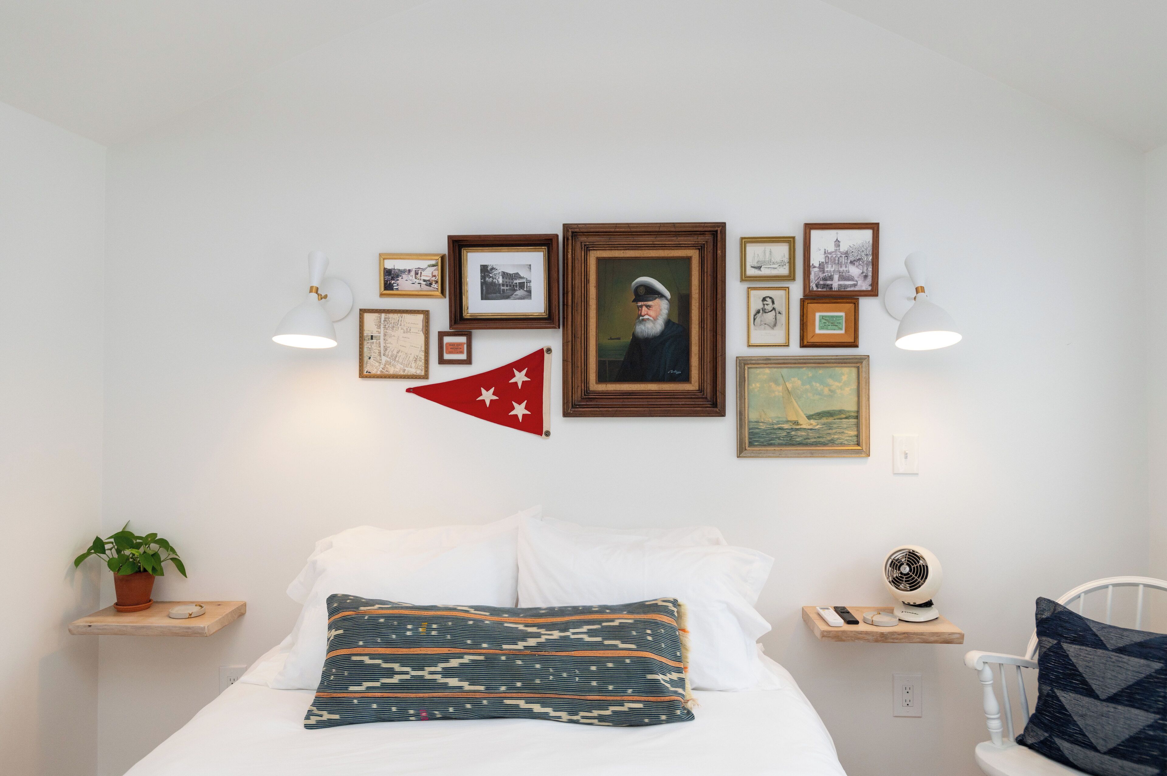 Un dormitorio ordenado con una cama, almohadas, una mesita de noche, una lámpara, un ventilador y una pared decorada con cuadros y una bandera.