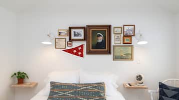 Un dormitorio ordenado con una cama, almohadas, una mesita de noche, una lámpara, un ventilador y una pared decorada con cuadros y una bandera.